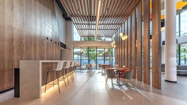 Un intérieur moderne avec un comptoir d’accueil en bois, un bar avec des tabourets et un espace repas doté de tables et de chaises rondes.