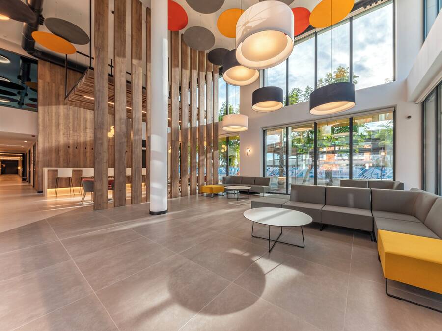 A spacious lobby with a modern design, featuring a large seating area, a staircase, and large windows offering views of the outdoors.