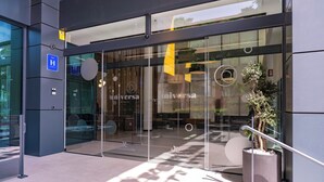 Une entrée d’hôtel moderne avec des portes en verre, un espace salon visible à l’intérieur et un petit jardinier à l’extérieur.