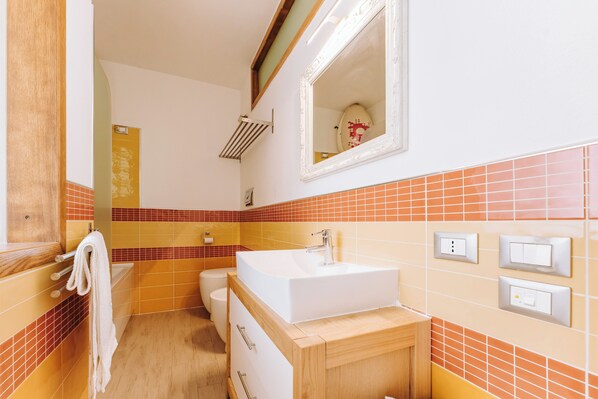 Un baño con lavamanos blanco, tocador de madera, paredes con azulejos naranja, un espejo y un perchero para toallas.