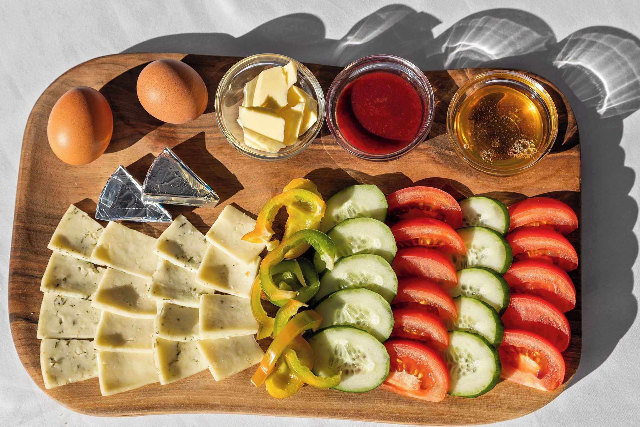 A wooden board with sliced tomatoes, cucumbers, bell peppers, eggs, butter, and a glass of juice.