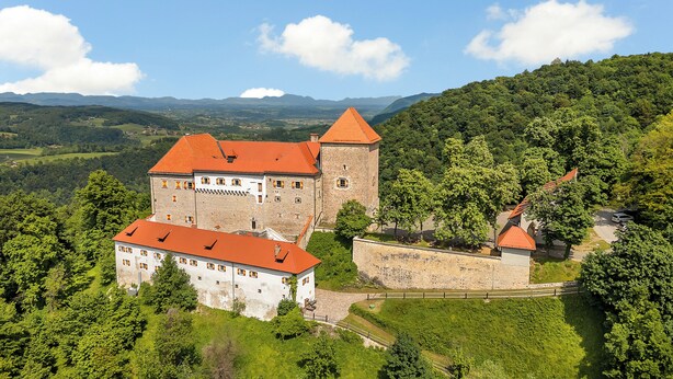 A castle with a red-tiled roof, surrounded by lush greenery and hills.