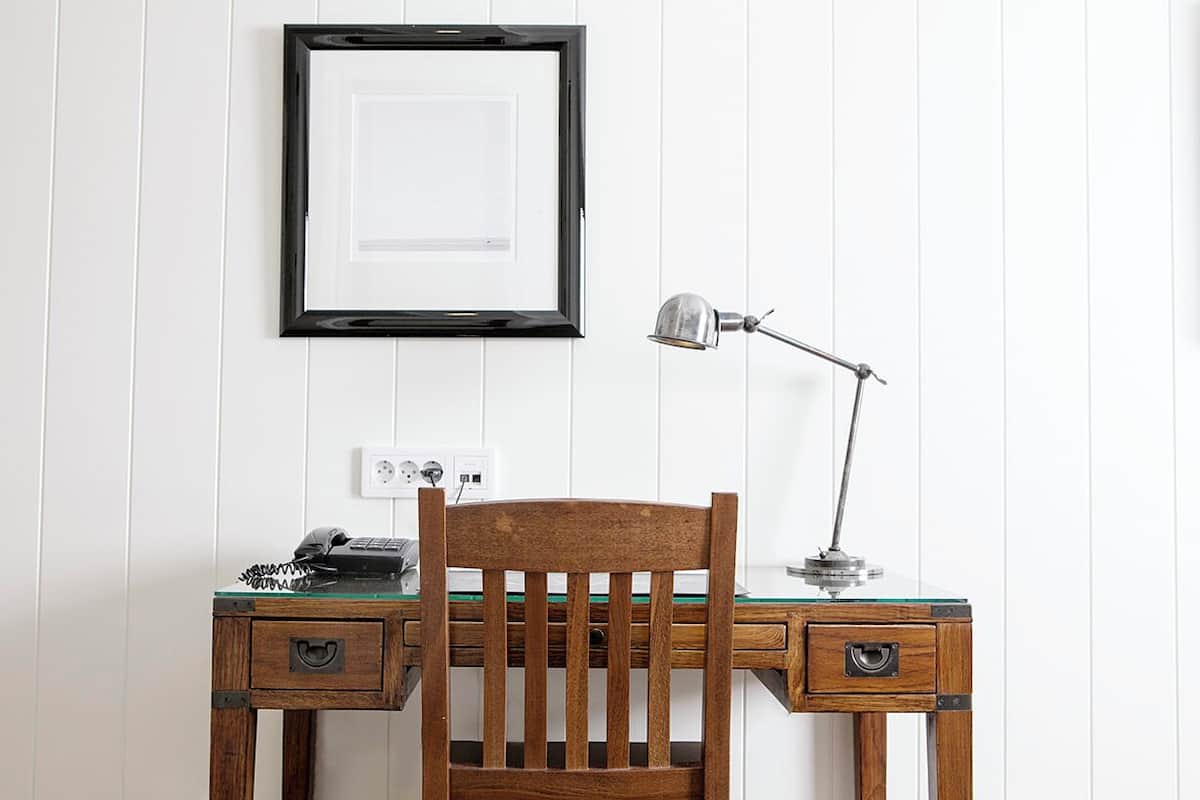 A wooden desk with a chair, a desk lamp, and a framed picture on the wall.