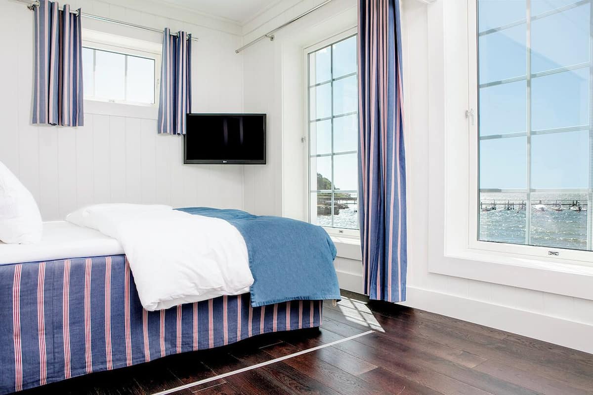 A hotel room with a bed, a TV mounted on the wall, and large windows offering a view of the water.