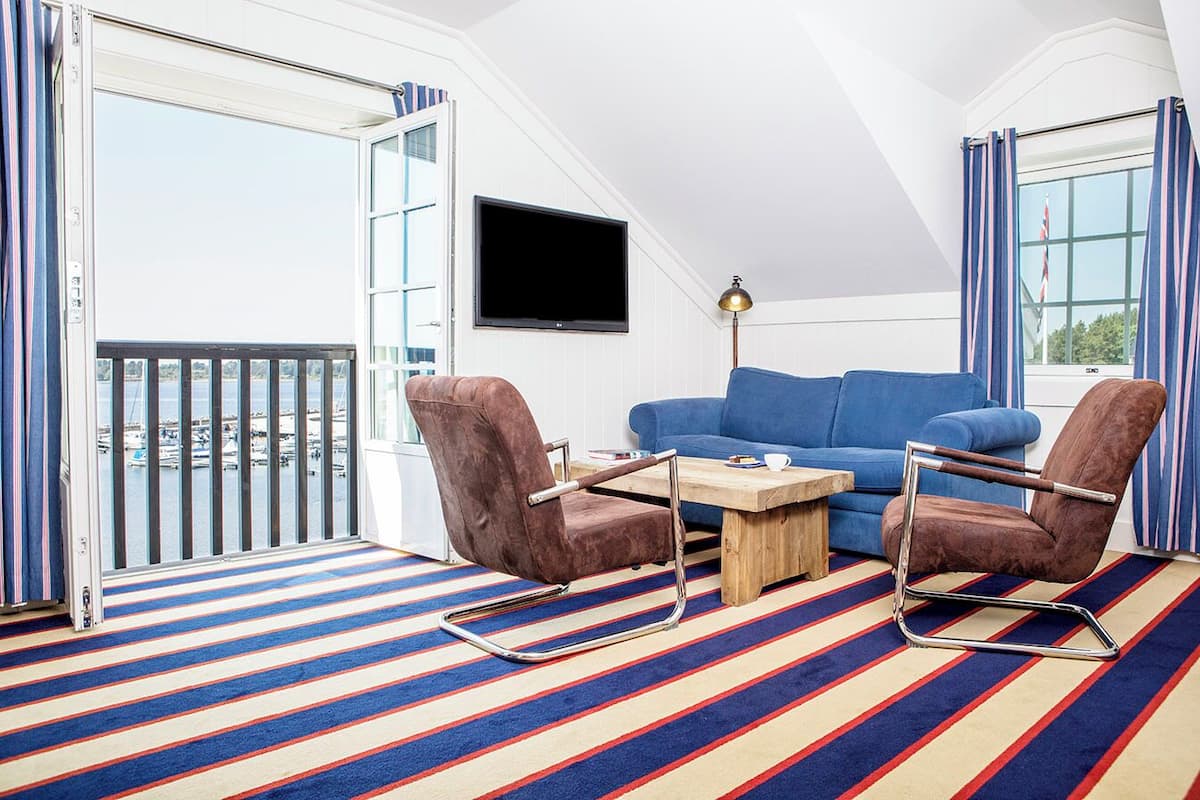 A room with a blue sofa, two brown chairs, a wooden coffee table, a TV mounted on the wall, and a balcony with a striped floor.