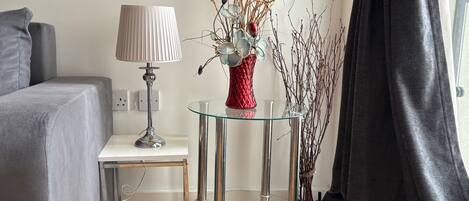 A modern living room with a grey sofa, a glass coffee table with a red vase, a lamp, and a small side table.