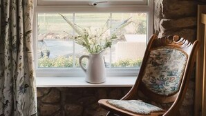 A room with a stone wall, a window with curtains, a white pitcher with flowers, and a wooden chair with floral upholstery. - Boutique Luxury at The Nook (Cockermouth)