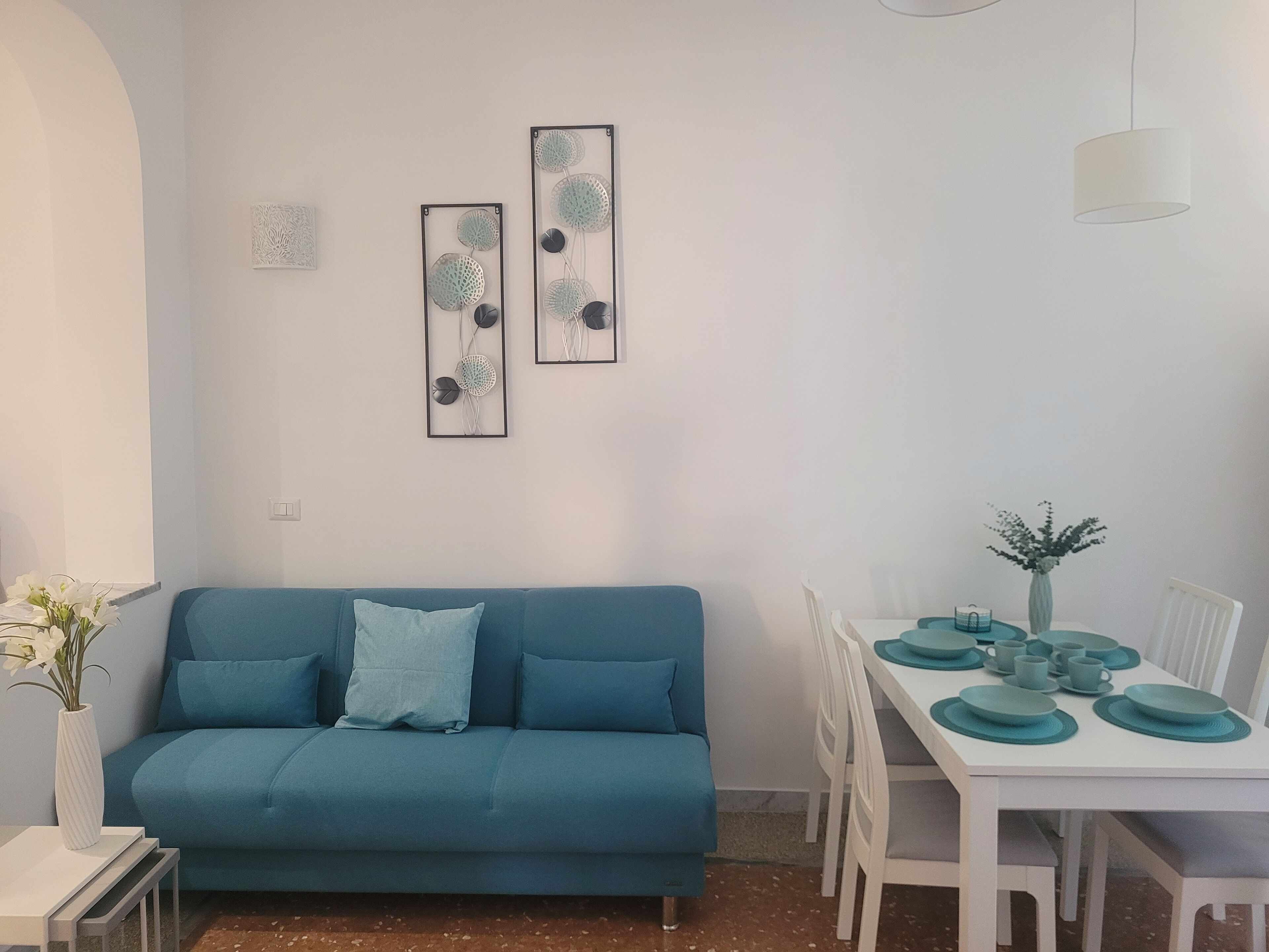 A modern living room with a blue sofa, a dining table set with teal plates, and two wall art pieces.