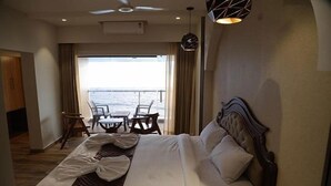 Una habitación de hotel con una cama grande, vista al mar, una zona de comedor con sillas y un ventilador de techo.