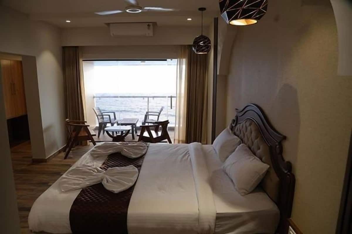 A hotel room with a large bed, a view of the sea, a dining area with chairs, and a ceiling fan.