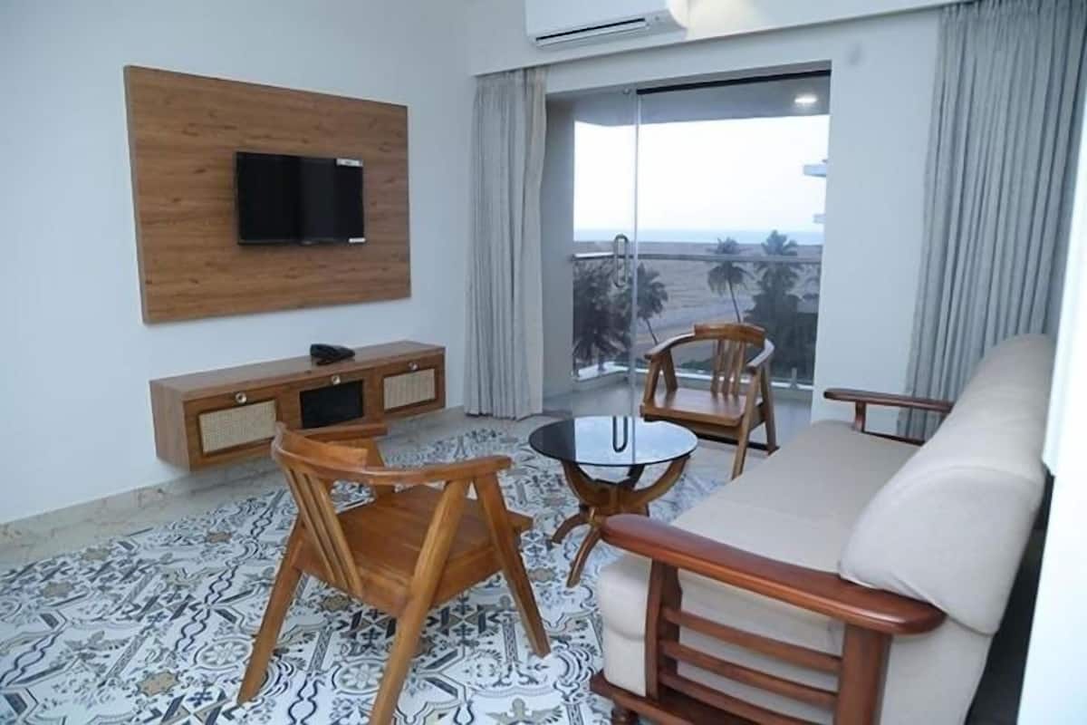 A modern living room with a sofa, two wooden chairs, a small round table, a TV mounted on a wooden panel, and a balcony with a view.
