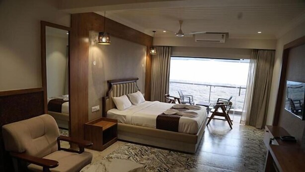 Amplia habitación de hotel con una cama grande, una zona de estar con una silla y una mesita pequeña, un ventanal con vista al mar y un ventilador de techo.