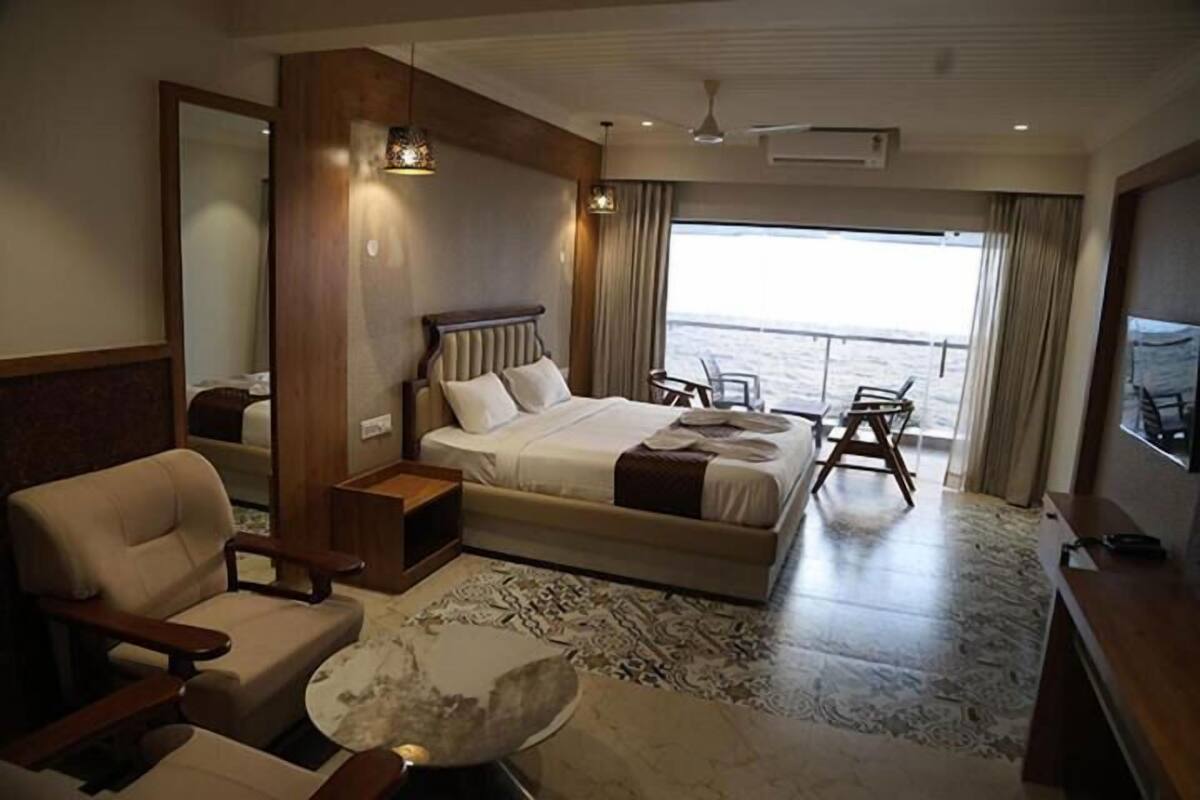 A spacious hotel room with a large bed, a seating area with a chair and a small table, a large window with a view of the sea, and a ceiling fan.