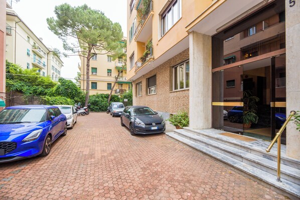 A paved driveway with parked cars, a building entrance with a glass door, and a staircase leading up to the entrance. - Spacious flat with Balcony (Genoa)
