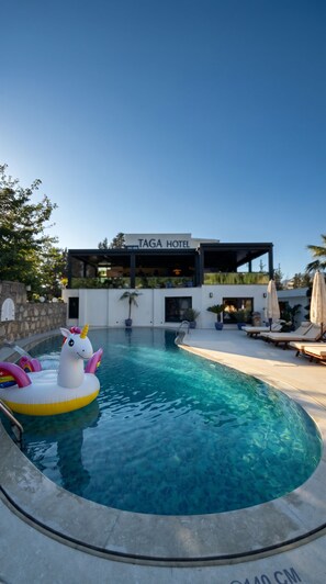 Una piscina con un flotador de unicornio, un edificio moderno con el letrero 'TAGA HOTEL' al fondo, y sillones de descanso a lo largo de la piscina.
