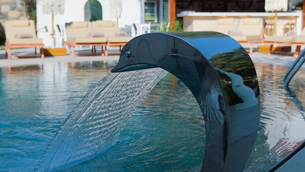 Une piscine agrémentée d’une fontaine en forme de dauphin, de chaises longues et d’une structure à toit de chaume en arrière-plan.