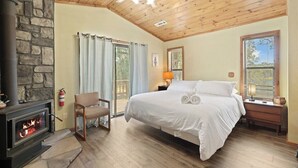 Une chambre avec une cheminée en pierre, un lit, une chaise et une fenêtre avec des stores.