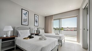 Ein Hotelzimmer mit zwei Betten, einem Nachttisch mit Lampe, einem gerahmten Bild und einem Balkon mit Aussicht.