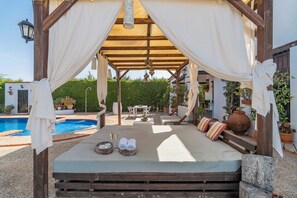 A cozy outdoor bed with a canopy, a pool, and a dining area in the background. - Holiday Home Carula (La Puebla de Cazalla)