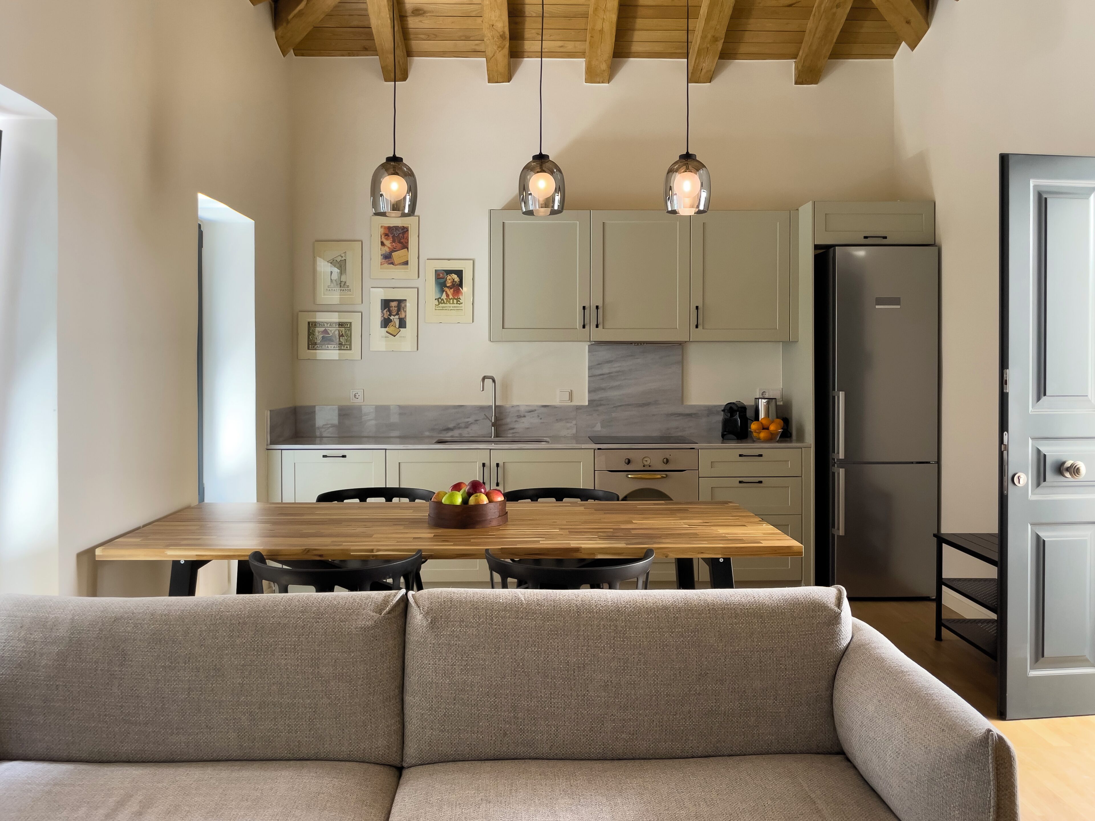 Een moderne keuken met een houten eettafel, een inbouwoven en een koelkast. Er hangen afbeeldingen in lijsten aan de muur en staat een schaal met fruit op de tafel.