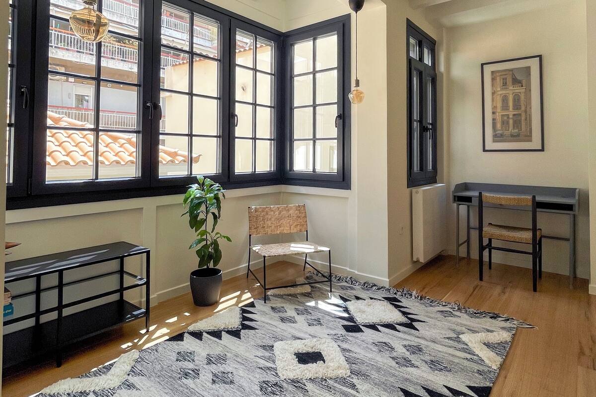 A spacious room with large windows, a black and white patterned rug, a wooden floor, a chair, a small table, and a potted plant.
