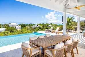 A poolside area with a wooden table and wicker chairs, overlooking a clear blue pool and a tropical landscape.