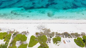 Vue aérienne d’une plage aux eaux turquoise et au sable blanc. On y aperçoit des zones de végétation verdoyante et quelques bateaux sur le rivage.