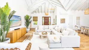 A spacious living room with a high ceiling, white sofas, a wooden coffee table, and a large painting of a beach scene. - AMANERA "peaceful water" NEW Villa with Ocean View - Walk to Beach (The Bight Settlement)
