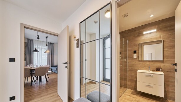 A modern hotel room with a wooden floor, a glass partition, a bathroom with a wooden vanity, and a dining area with a table and chairs.