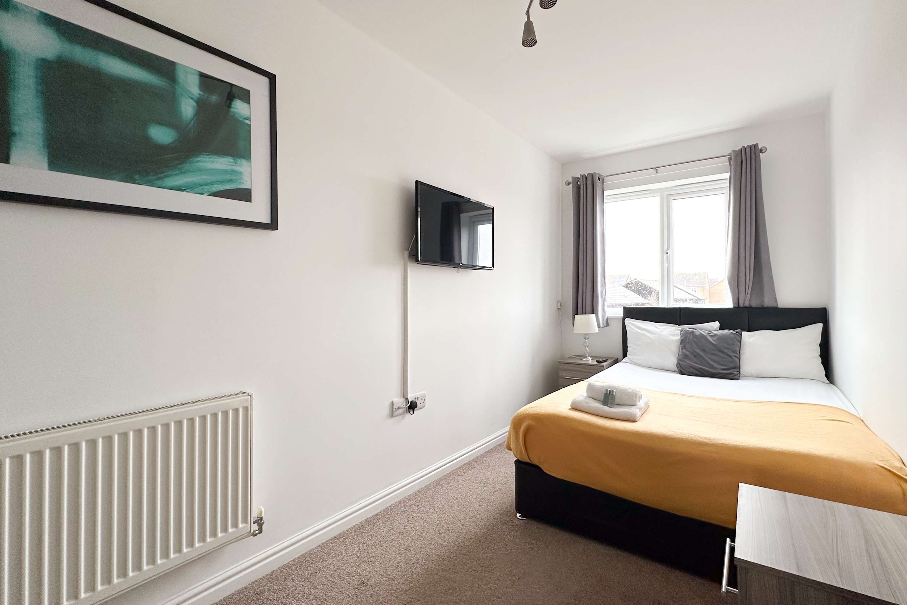 Een hotelkamer met een bed, een aan de muur bevestigde televisie, een raam met gordijnen, een radiator en een ingelijst schilderij.