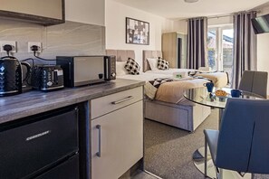 Kompakti hotellihuone, jossa on pieni keittiönurkkaus, kuviollisilla tyynyillä varustettu sänky, lasipöytä ja näkymä ulos.