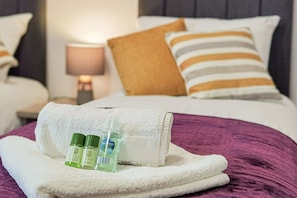 Een net opgemaakt bed met een witte handdoek, twee flessen Eden douchegel en een paars deken.