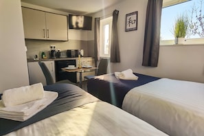 Een kleine hotelkamer met twee bedden, een kitchenette, een eettafel en een raam met gordijnen.