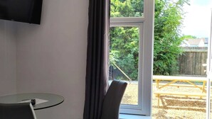 Een kamer met een glazen tafel, een stoel, een bed, een aan de muur bevestigde televisie en een raam met uitzicht op een tuin en een houten terras.