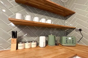 A kitchen counter with wooden shelves holding mugs, a coffee grinder, tea and sugar containers, a paper towel holder, and a green toaster.