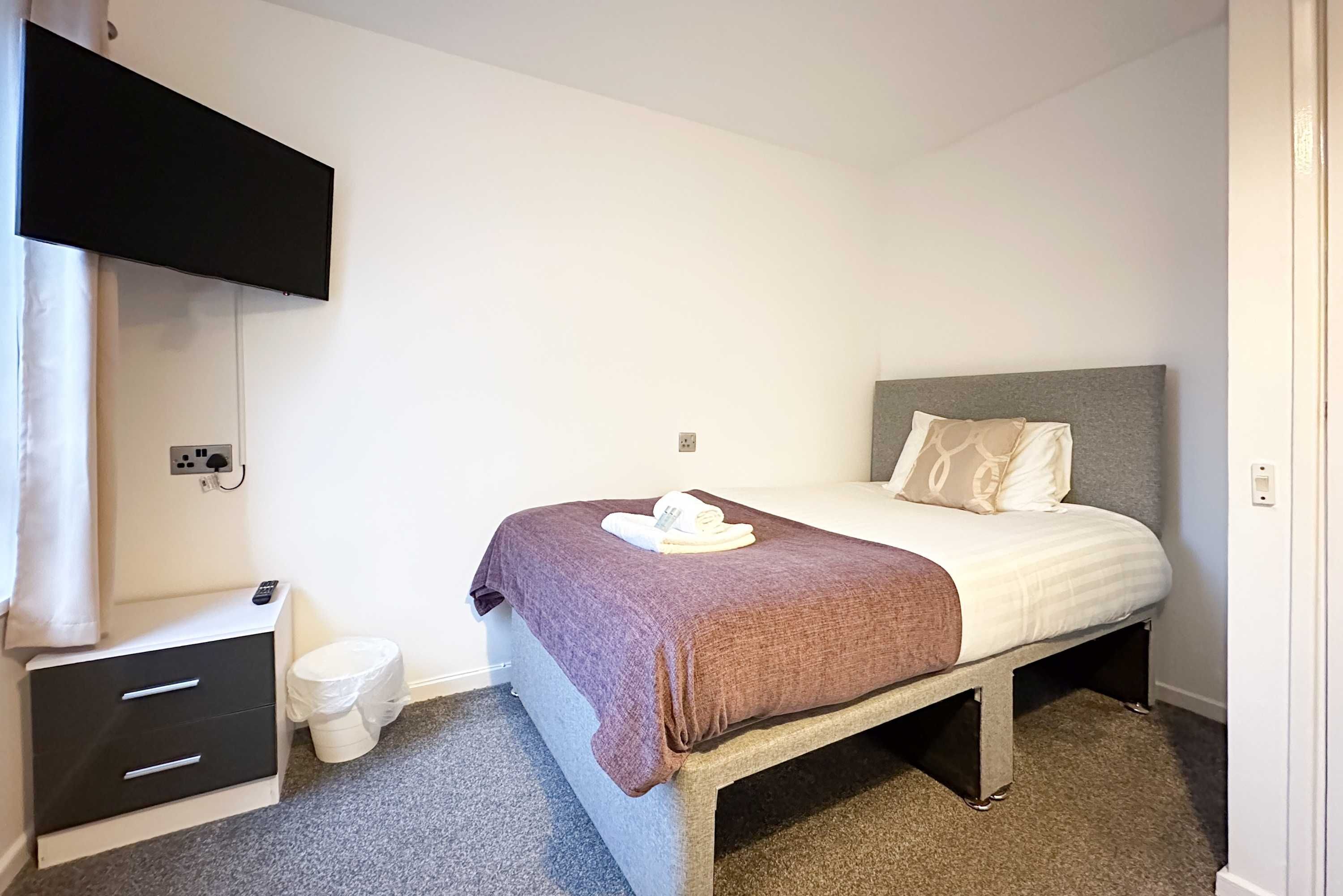 A hotel room with a bed, a bedside table with a remote control, a small drawer unit, a toilet, and a wall-mounted TV.