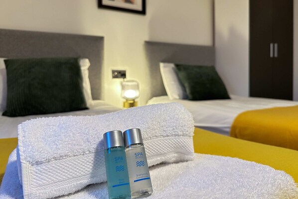 A hotel room with a neatly made bed, two bottles of body wash and conditioner, and a night lamp on the bedside table.
