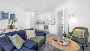 Una sala moderna con un sofá azul, un sillón verde y una mesa de centro de madera. Se ve una cocina al fondo, con armarios blancos y un comedor con una mesa y sillas.