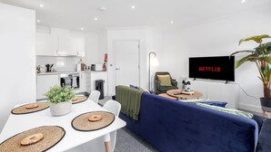 Una sala moderna con un sofá azul, una mesa de comedor con individuales, un televisor mostrando 'NETFLIX' y una cocina al fondo.