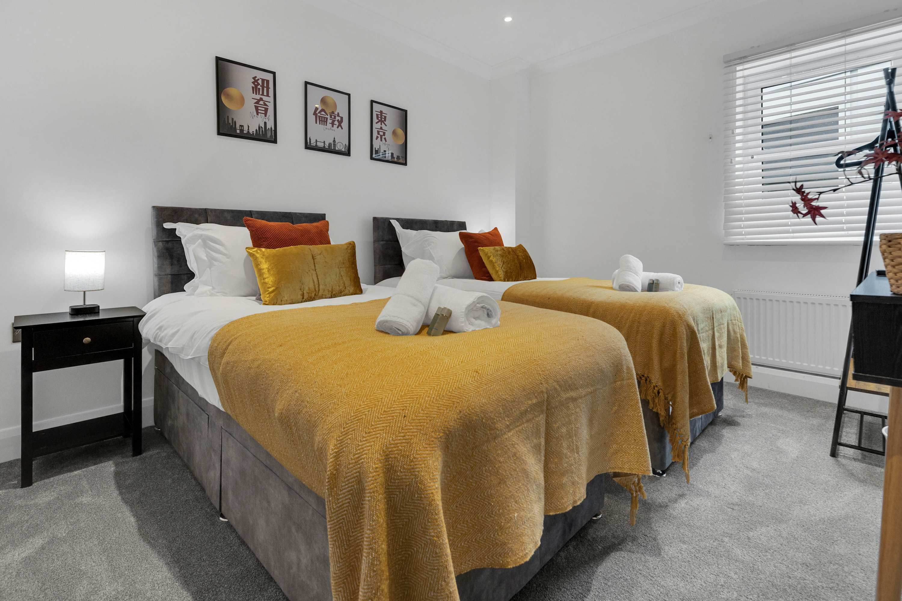 A hotel room with two beds, a bedside table, a lamp, and a window with blinds.