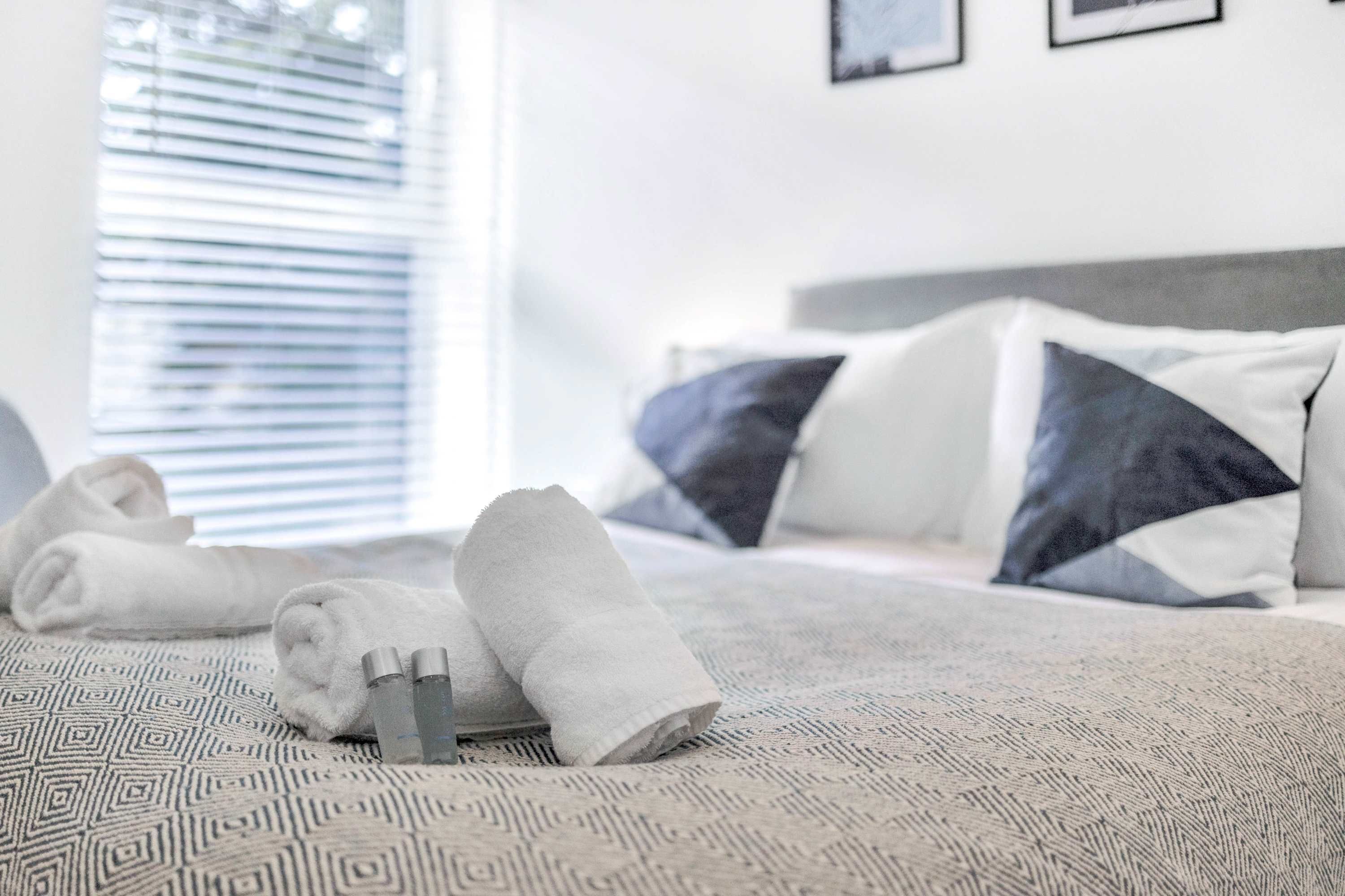 A neatly made bed with rolled towels and toiletries, a patterned bedspread, and a window with blinds.