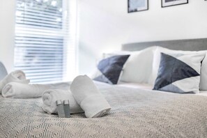 Een net opgemaakt bed met opgerolde handdoeken en toiletartikelen, een bed met een dessin en een raam met zonwering.