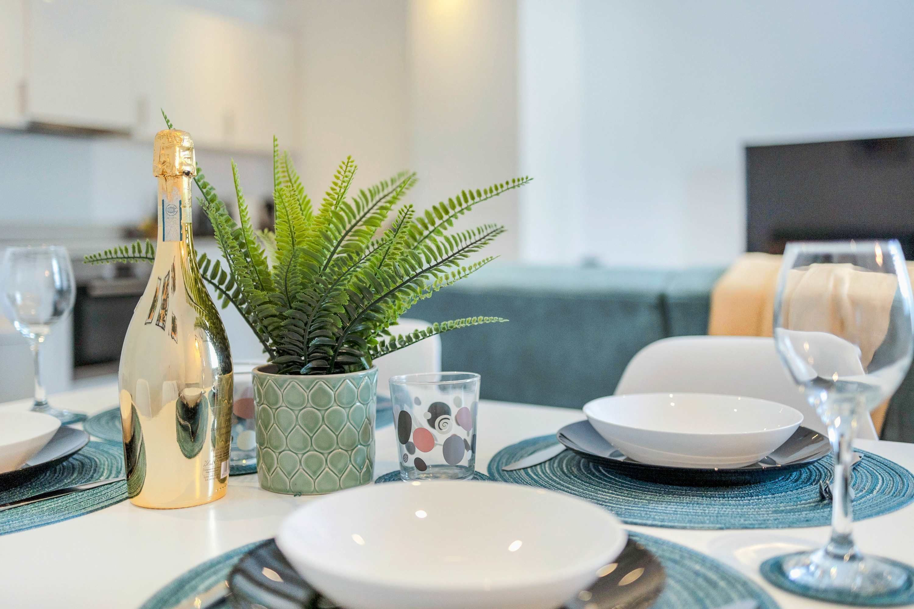 A dining table set with plates, a wine glass, a bottle of champagne, a decorative plant, and a patterned coaster.