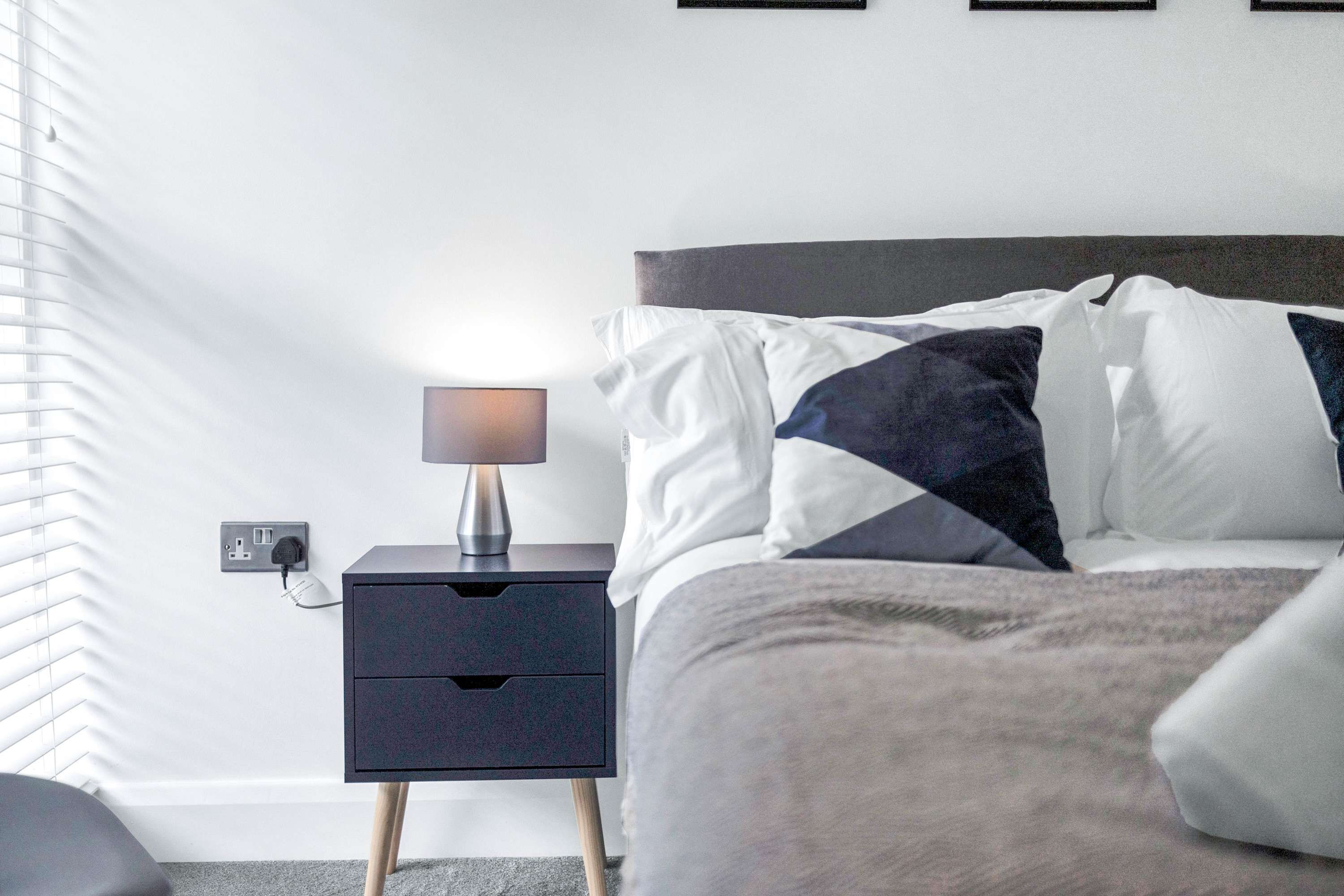 A neatly made bed with a dark headboard, a bedside table with a lamp, and a power outlet on the wall.