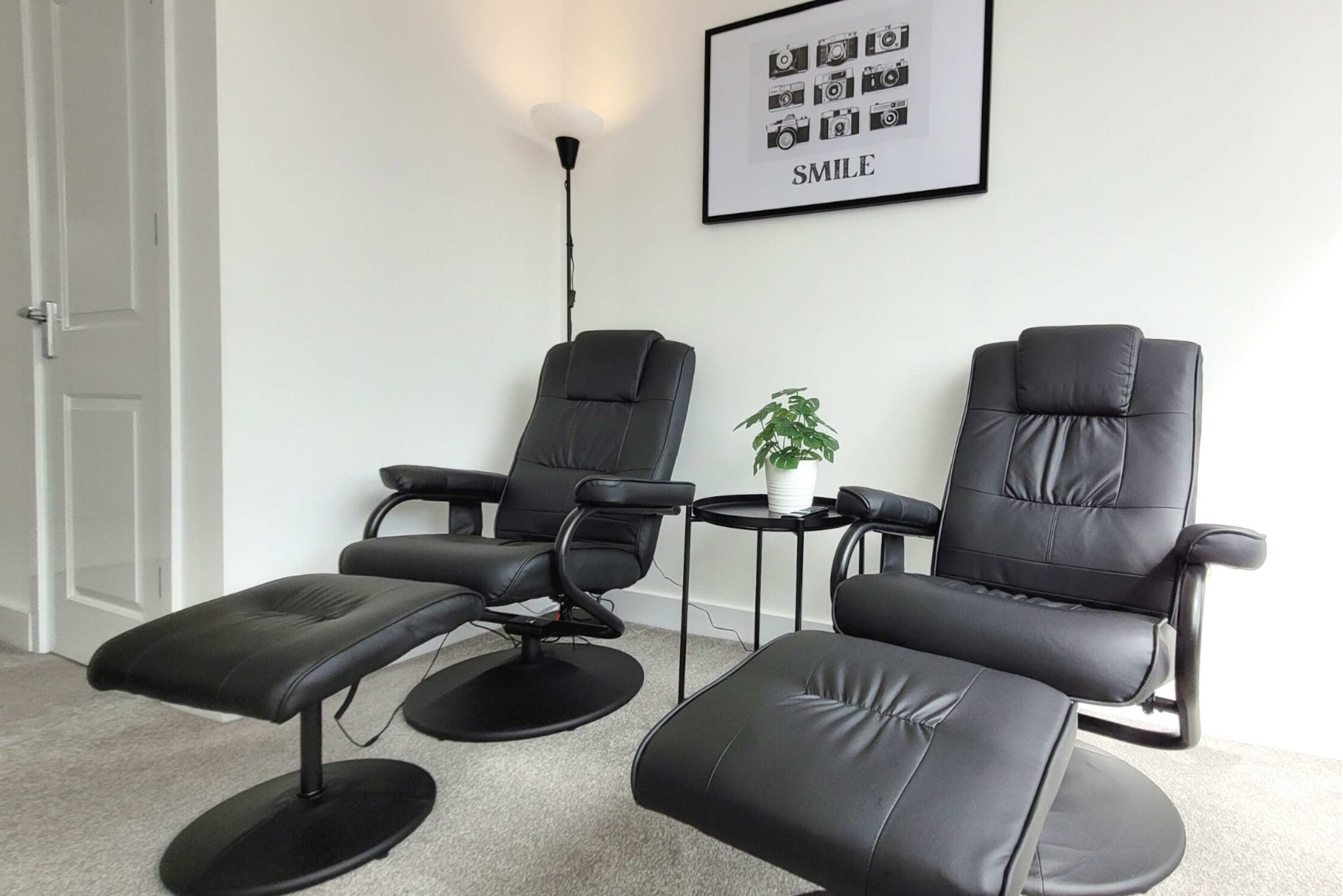 A room with three black leather chairs, a small white table with a plant, and a framed picture on the wall.