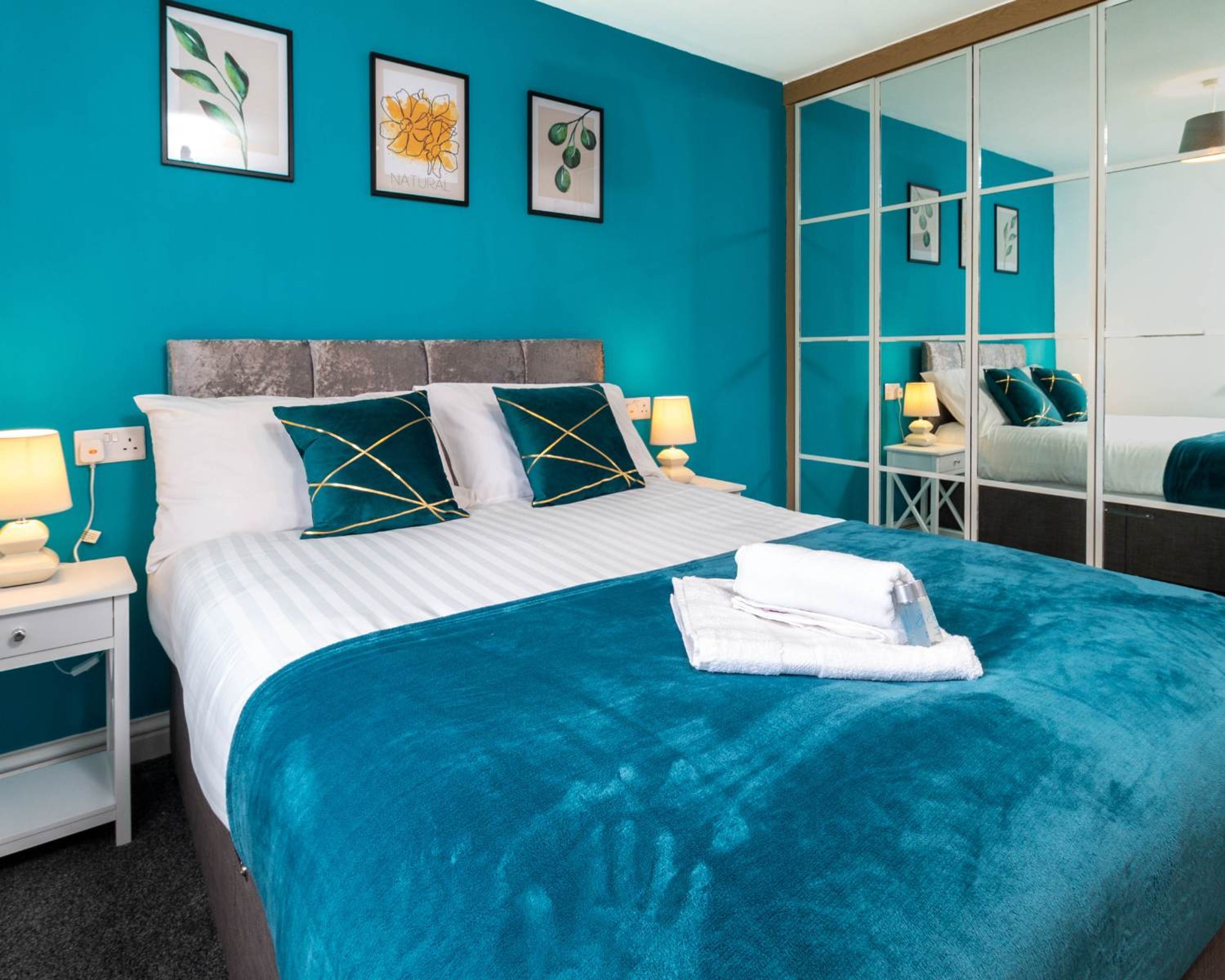 A hotel room with a large bed, teal walls, and a large mirror. There are two bedside tables with lamps and a small nightstand.