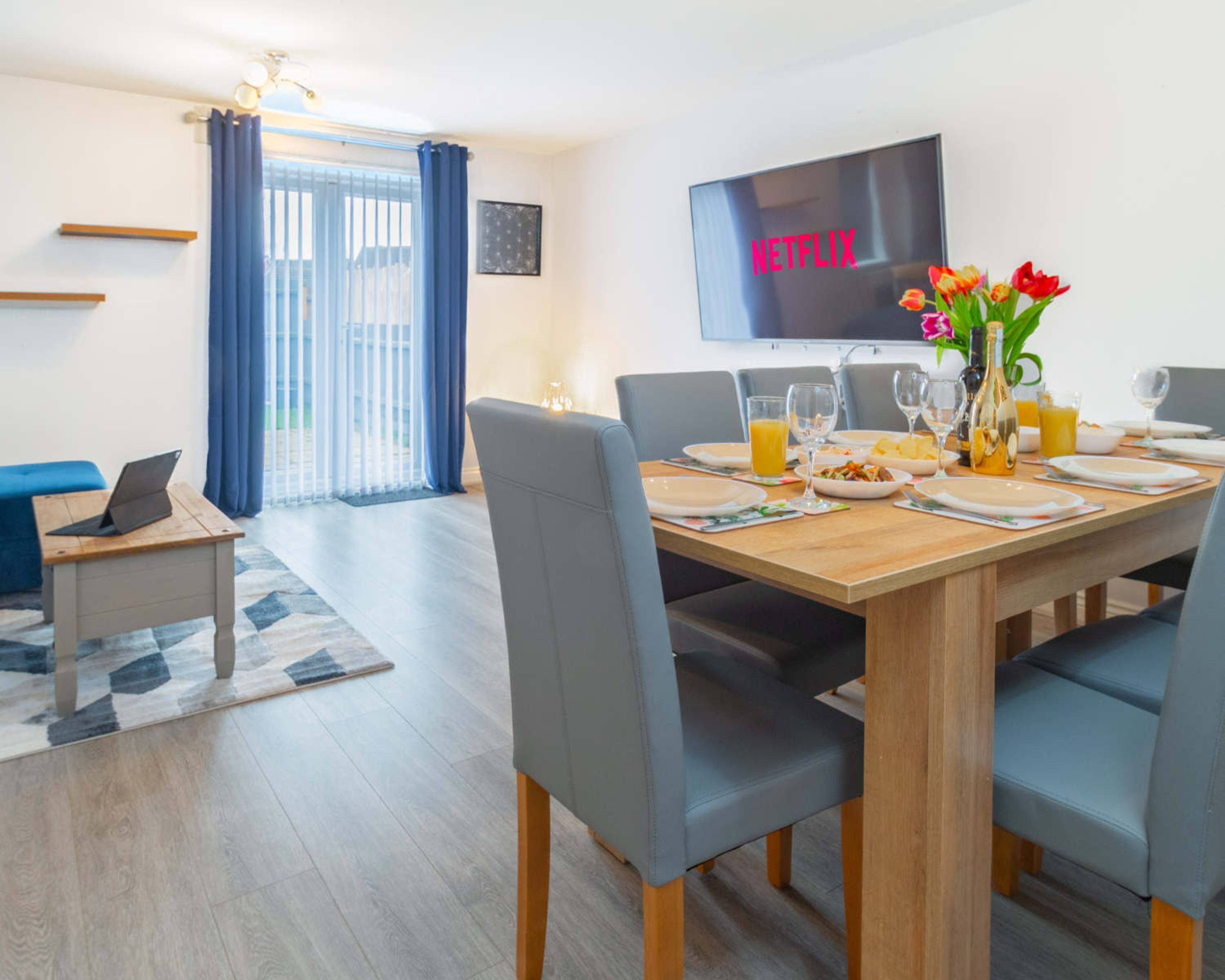 A dining area with a wooden table set for four, a laptop on a side table, and a TV mounted on the wall displaying "NETFLIX".