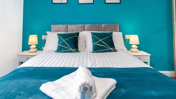 A hotel room with a teal wall, a bed with striped bedding, and two nightstands with lamps. There are three framed pictures on the wall above the bed.