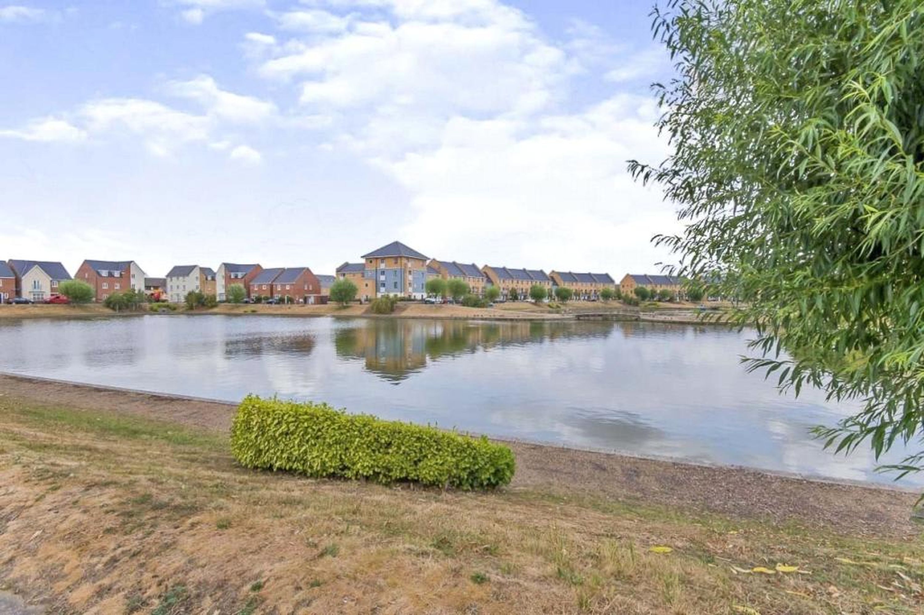 A residential area with houses along a calm water body, a well-maintained hedge, and a clear sky.