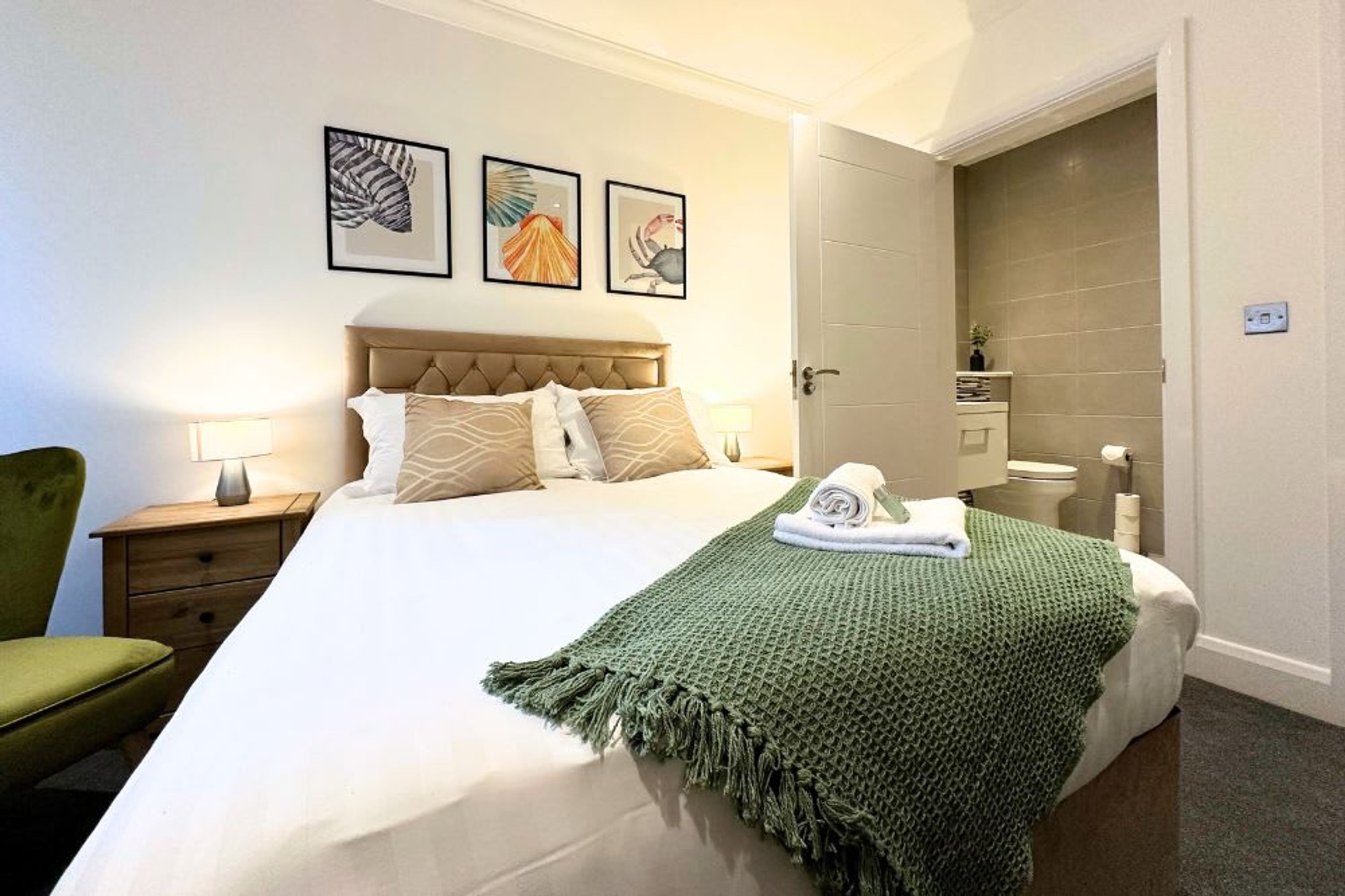 A neatly made bed with a green blanket, a headboard, and three framed artworks on the wall.